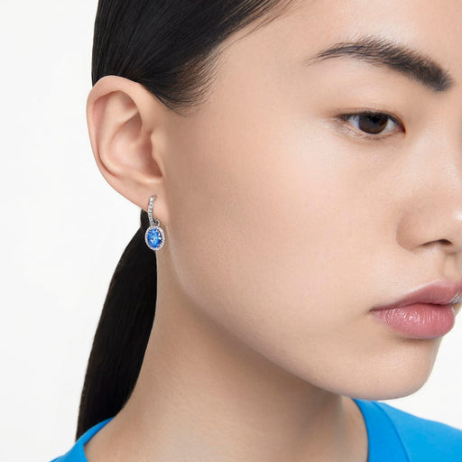 Close-up of a woman with sleek hair wearing silver-tone rhodium-plated drop earrings with oval-cut blue crystal stones and cubic zirconia halos, complementing her blue top and providing a touch of elegance.